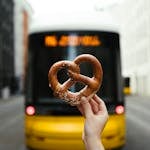 Hand holding pretzel in front of a Berlin street with a yellow bus, capturing urban lifestyle.
