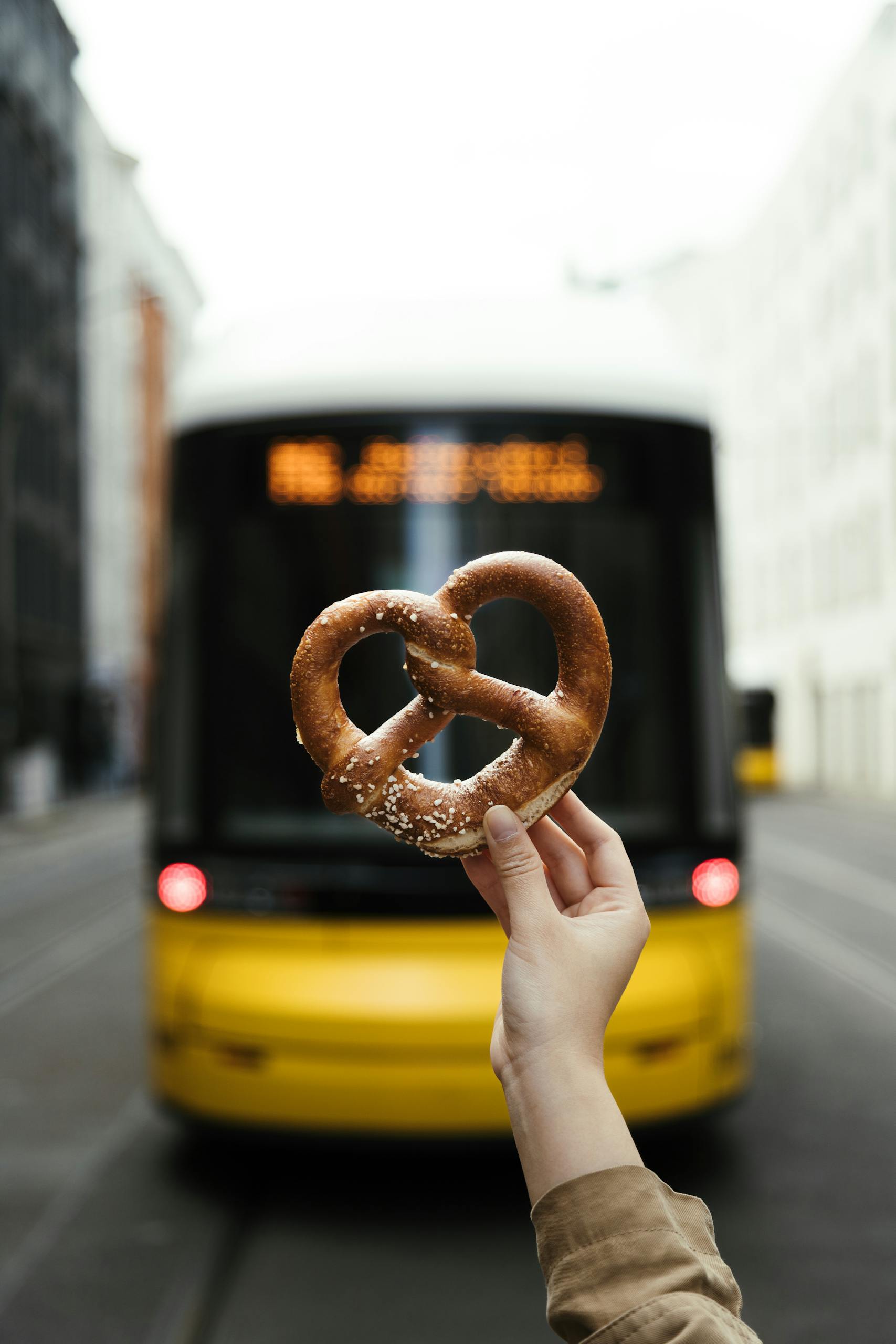 Hand holding pretzel in front of a Berlin street with a yellow bus, capturing urban lifestyle.